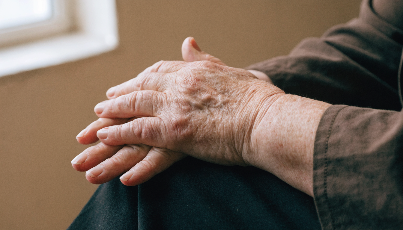 An older woman's hands resting one over the other in her lap by a window